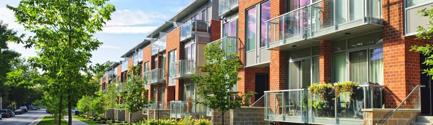 Moderne Reihenhausanlage mit roten Klinkerfassaden, Glasbalkonen und begrüntem Gehweg mit Bäumen.