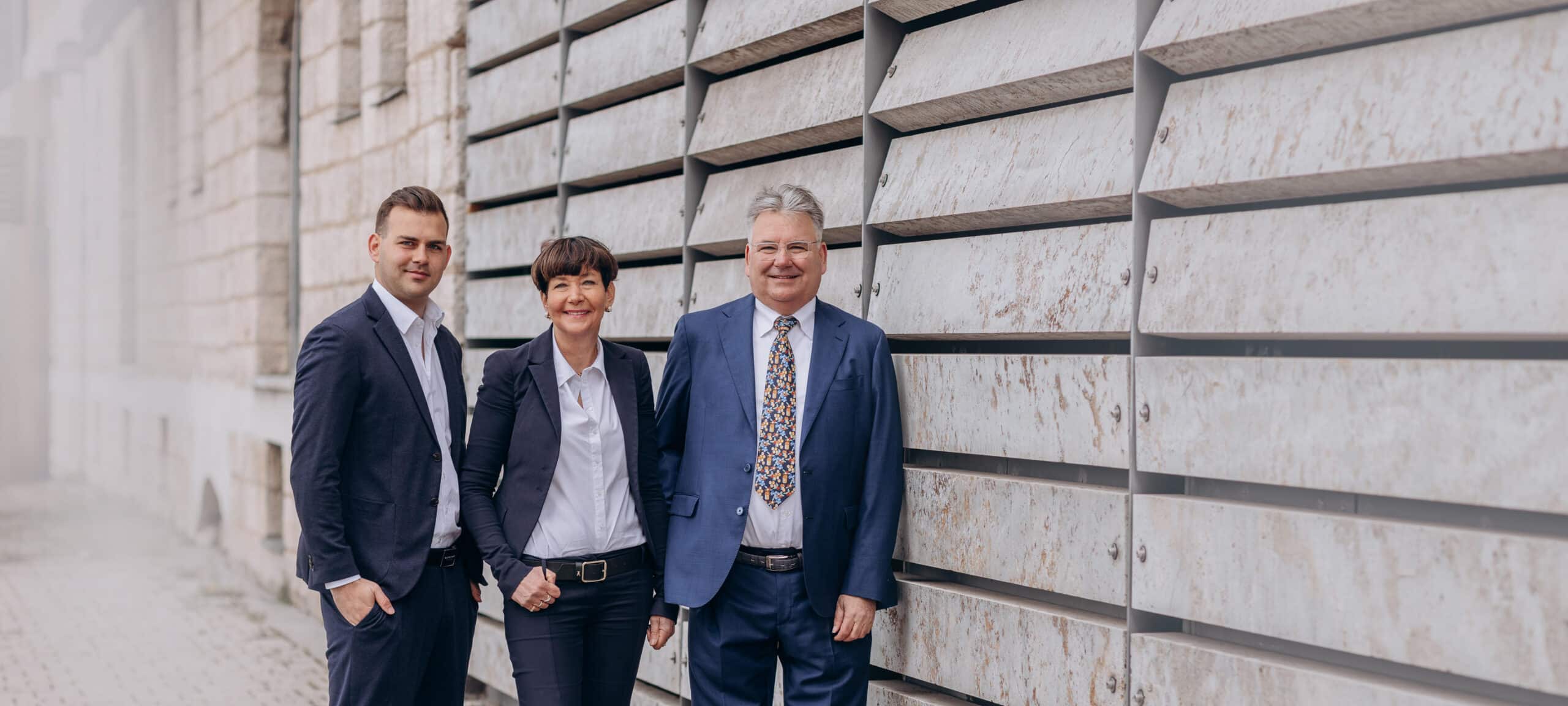 Zwei Männer und eine Frau in dunkler Businesskleidung stehen in die Kamera lächelnd nebeneinander auf einem gepflasterten Weg vor einer großflächigen, waagerecht strukturierten Metallfassade mit Natursteinoptik.
