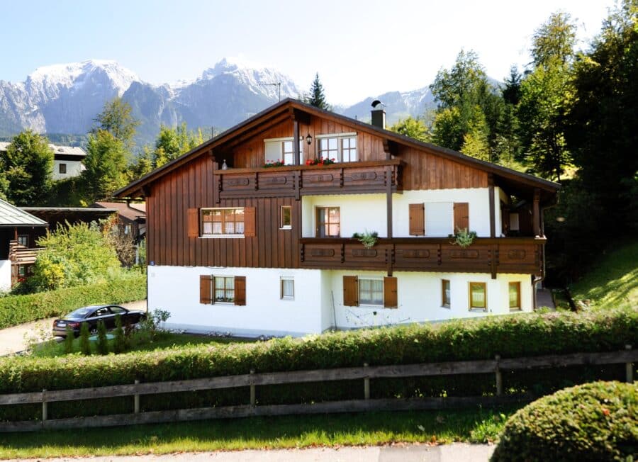 Zweigeschossiges Mehrfamilienhaus mit Holzfassade und Satteldach, Balkon und Fensterläden, im Vordergrund ein Holzzaun; Hintergrund zeigt bewaldete Berge mit Schnee.