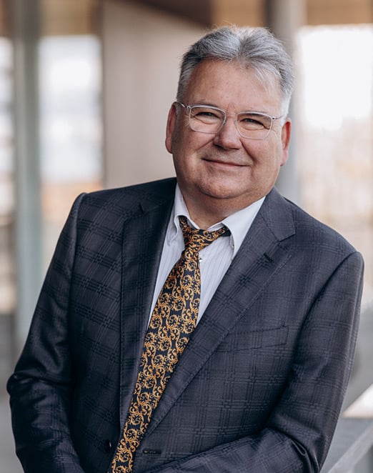 Ein Mann mit grauen Haaren, Brille, dunklem karierten Anzug, weißem Hemd und gold-braun gemusterter Krawatte steht leicht schräg und lächelt; im Hintergrund unscharfe Gebäudeelemente mit Licht.