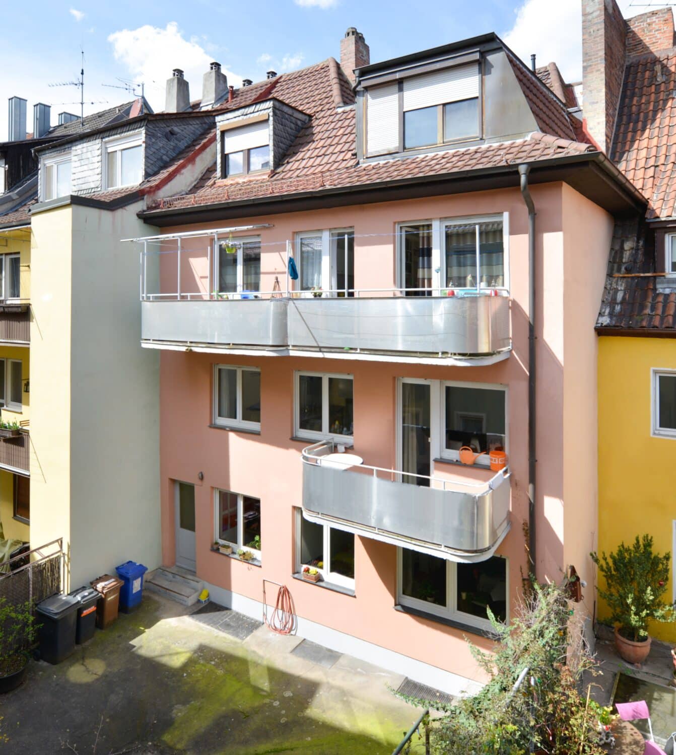Rückansicht eines gepflegten Mehrfamilienhauses mit rosafarbener Fassade, zwei Balkonen, Dachfenstern und Innenhof
