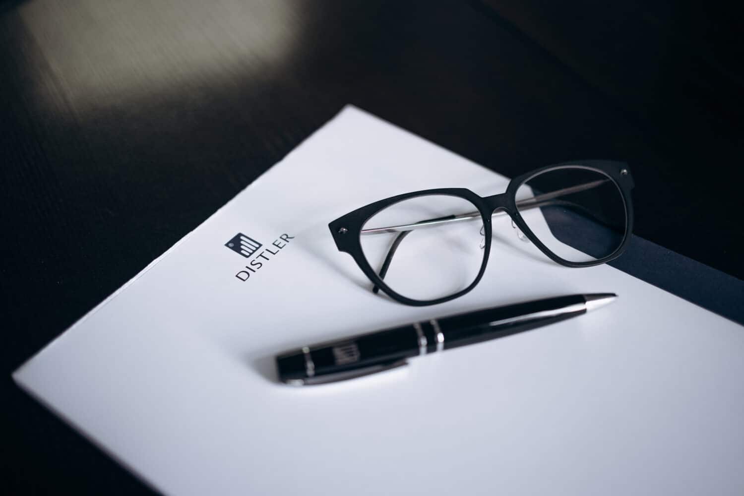 Schwarze Brille und schwarzer Kugelschreiber mit silbernen Details liegen auf einem weißen Schreibblock mit DISTLER-Logo, Hintergrund ist eine dunkle Tischoberfläche.
