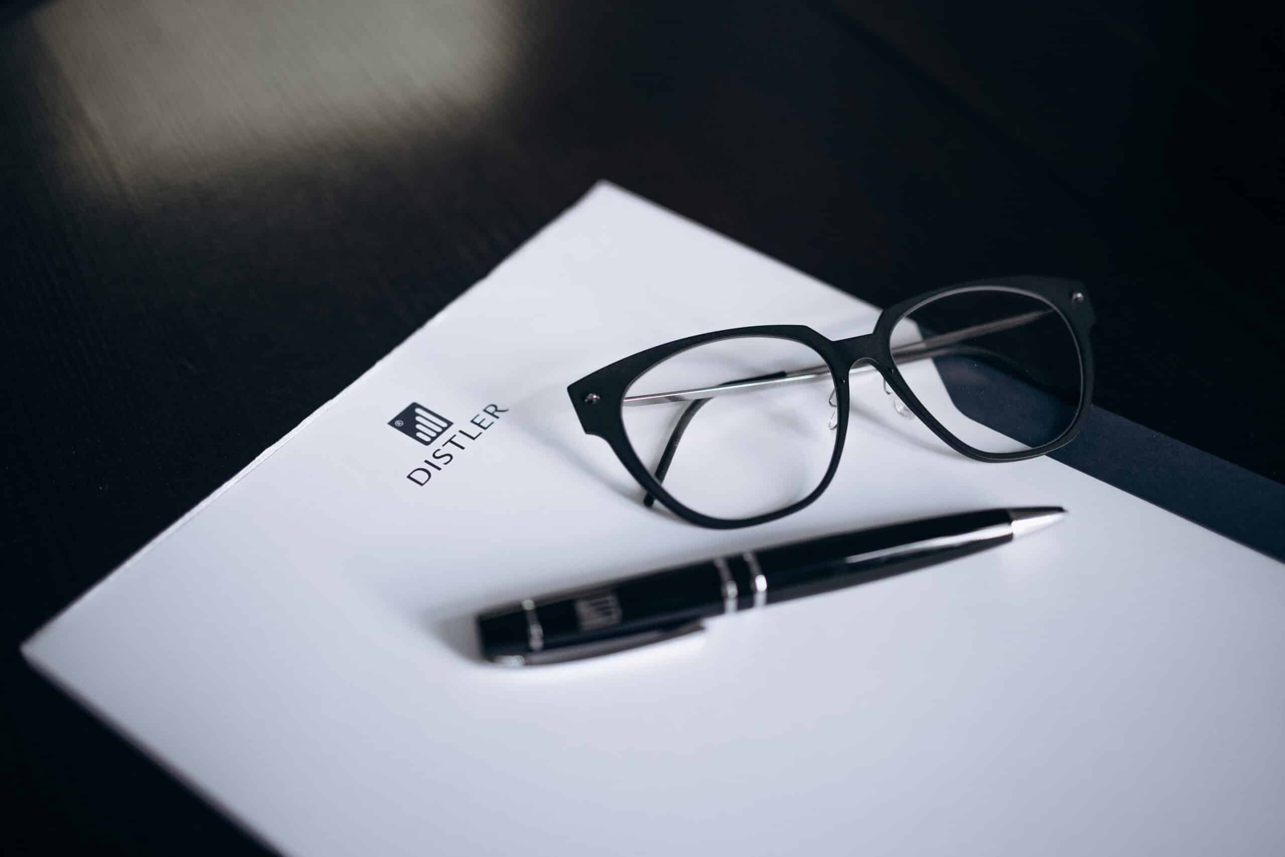 Schwarze Brille und schwarzer Kugelschreiber mit silbernen Details liegen auf einem weißen Schreibblock mit DISTLER-Logo, Hintergrund ist eine dunkle Tischoberfläche.