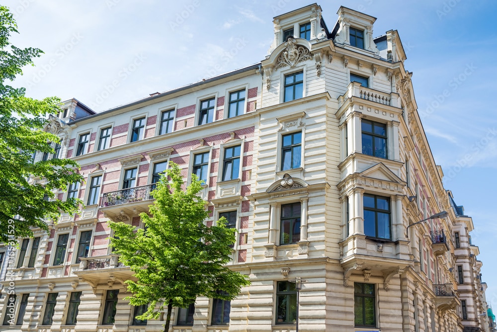 Viergeschossiges Wohn- und Geschäftshaus mit aufwendig verzierter Stuckfassade, Erkern, Balkonen und dekorativen Fensterelementen, im Vordergrund ein grüner Baum.