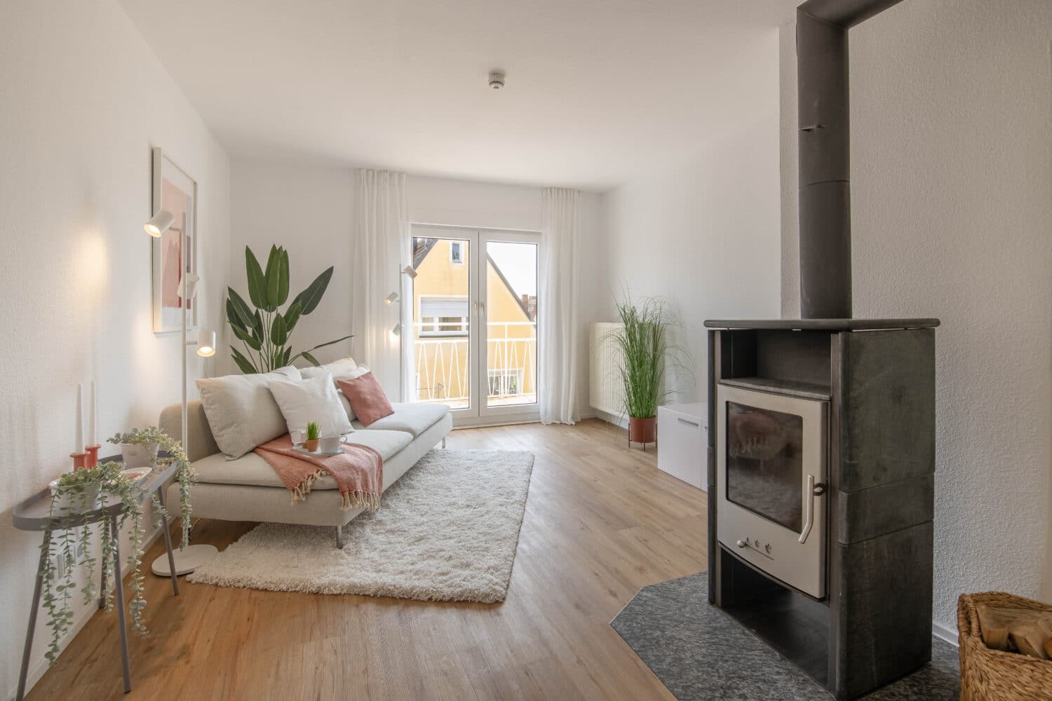 Modern eingerichtetes Wohnzimmer mit hellem Laminatboden, weißem Sofa mit Kissen und Decke, flauschigem Teppich, bodentiefen Fenstern mit Balkonzugang, Vorhängen, mehreren Zimmerpflanzen, einem Kaminofen im Vordergrund rechts und Tageslicht aus südlicher Richtung.