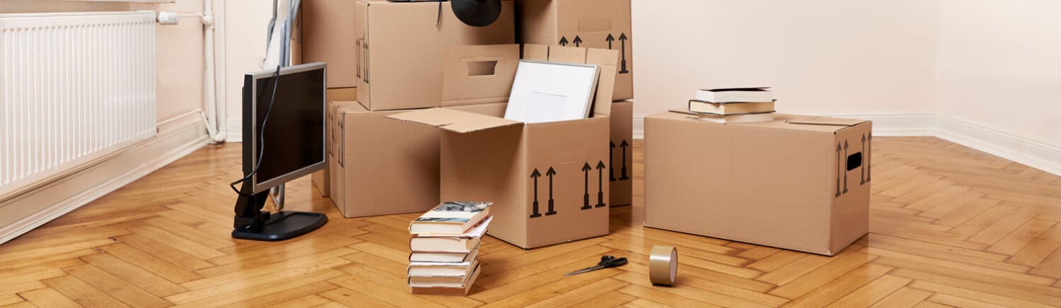 Umzugskartons, Bücher und Monitor auf Parkettboden in leerer Wohnung.