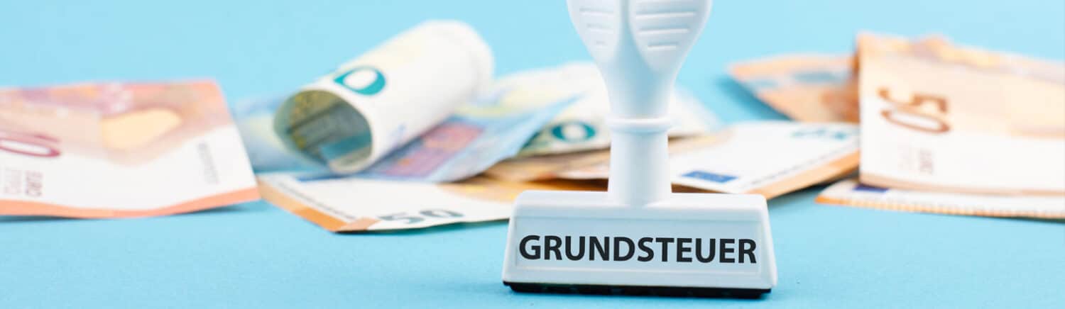 Stempel mit der Aufschrift „Grundsteuer“ liegt auf verstreuten Euro-Scheinen vor hellblauem Hintergrund.