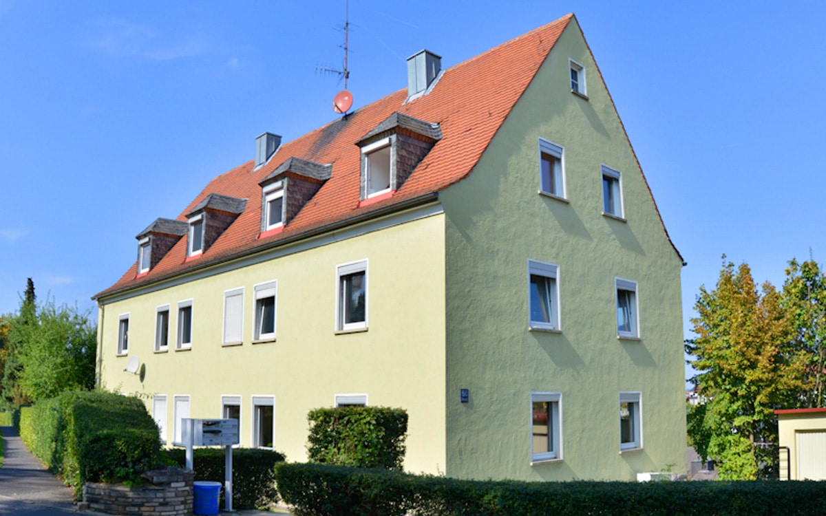 Dreigeschossiges Wohngebäude mit hellgelber Putzfassade, weißen Fensterrahmen und rotem Satteldach, davor ein niedriger grüner Strauch.