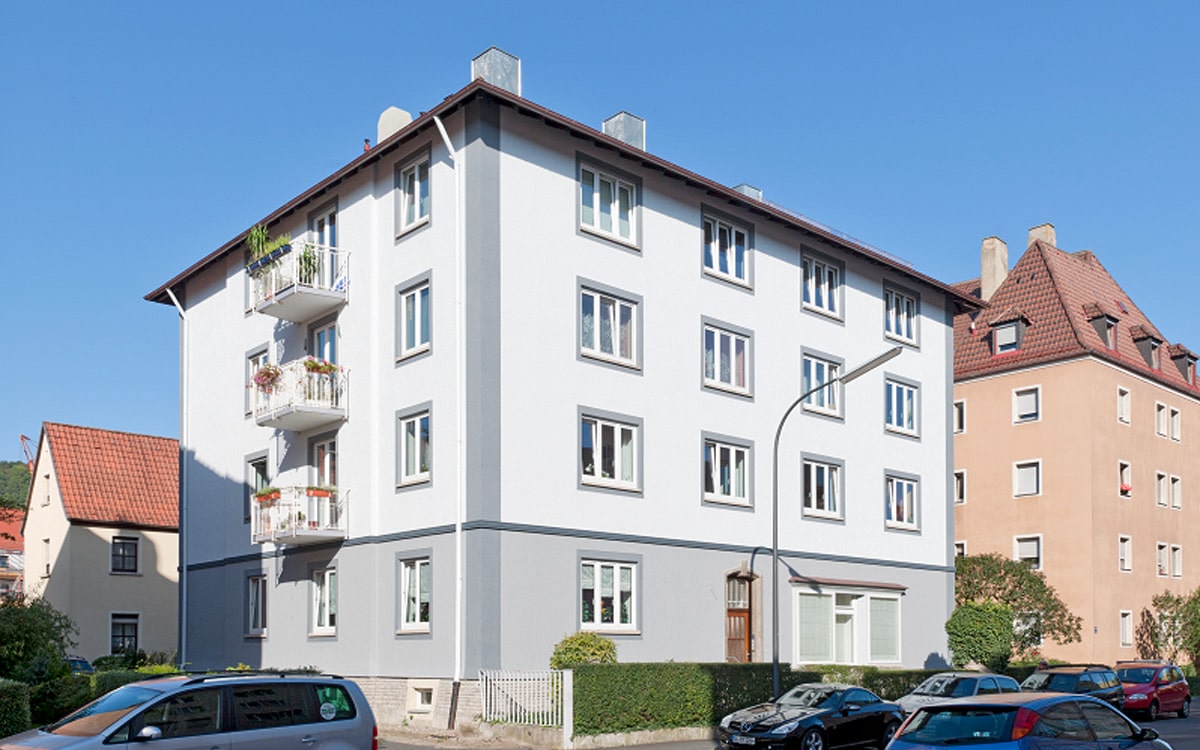 Helles, viergeschossiges Wohnhaus mit mehreren Balkonen, weißen Fenstern und hellgrauer Sockelzone, davor Gehweg und geparkte Fahrzeuge.