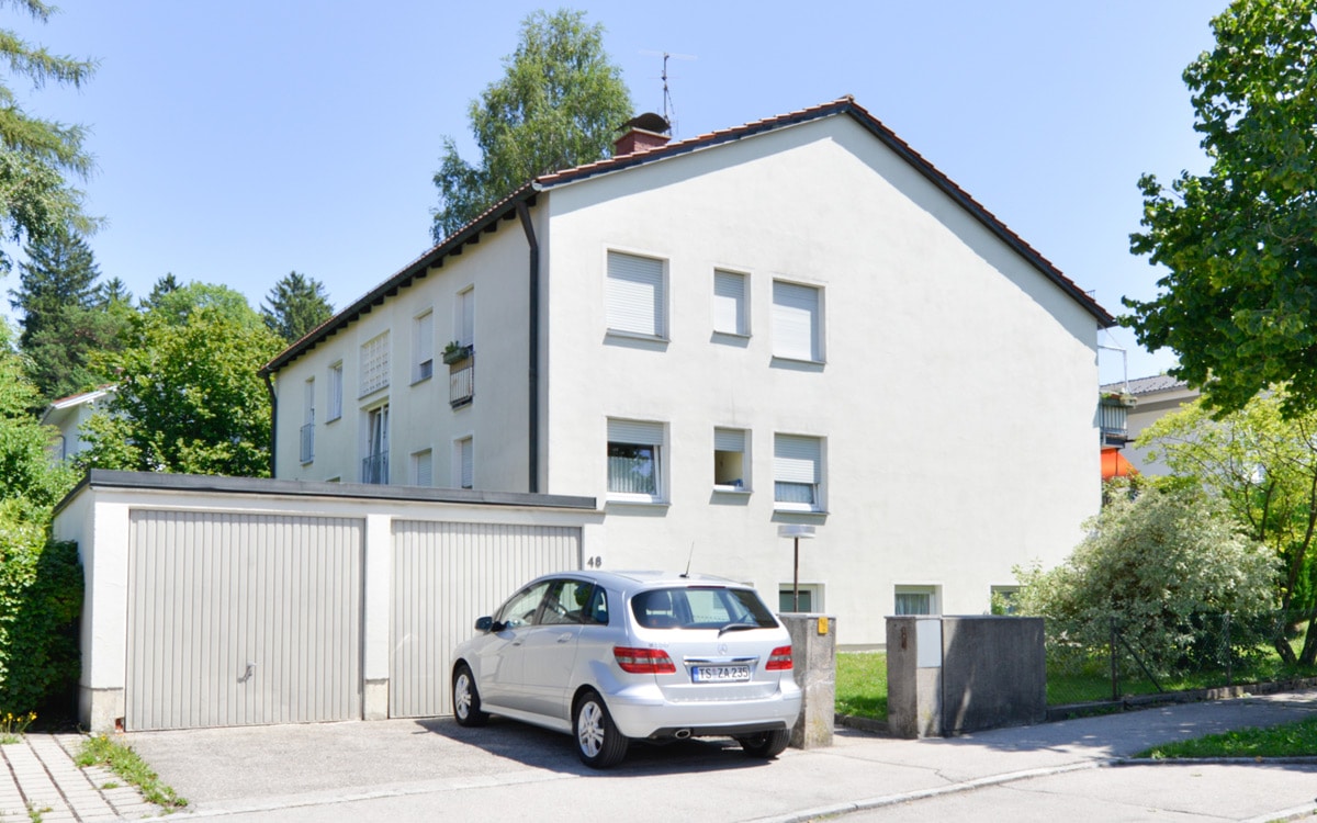 Rückansicht eines gepflegten Mehrfamilienhauses mit weißer Fassade, Garage, Parkplatz und Grünanlage.