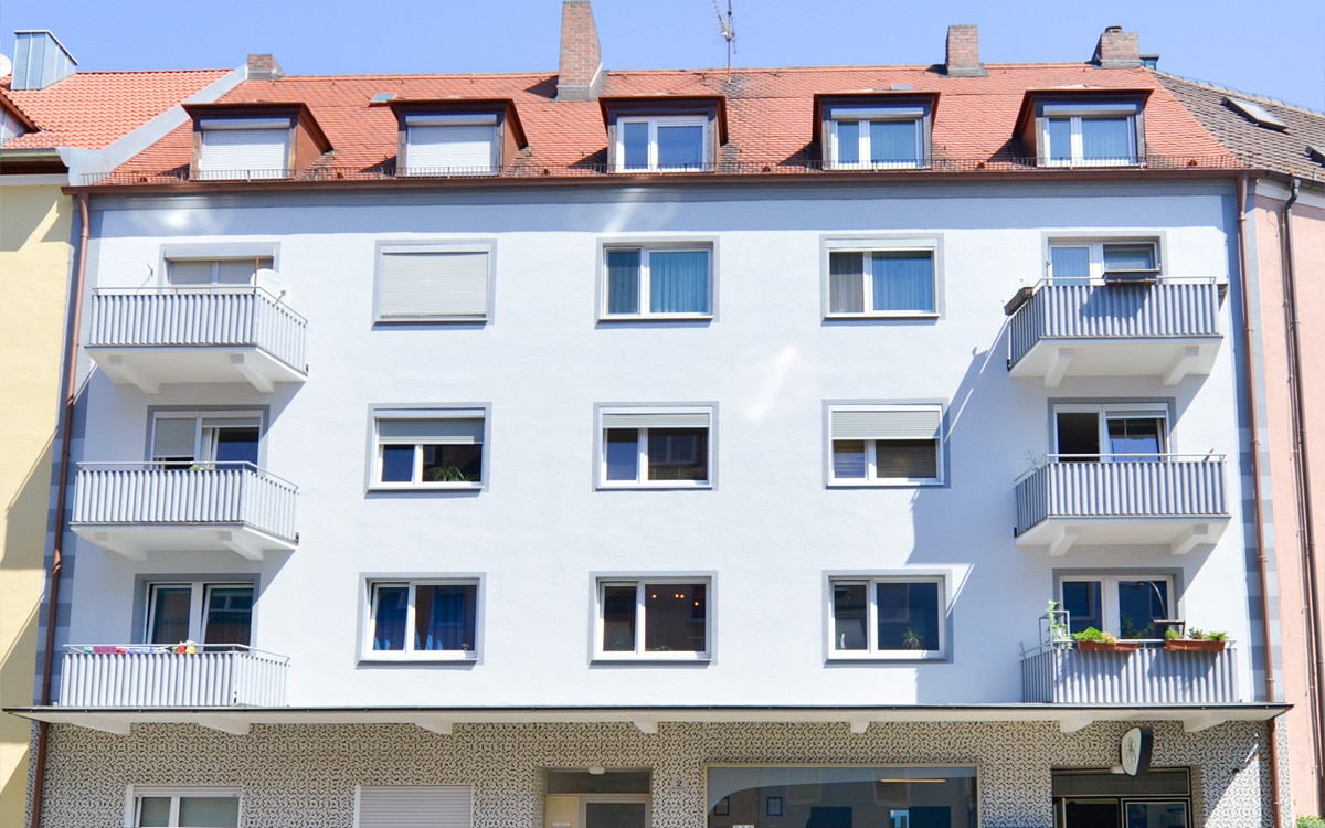 Viergeschossiges Gebäude mit weißer Fassade, mehreren Fenstern, einem Ladengeschäft im Erdgeschoss und Balkonen zur Straße.