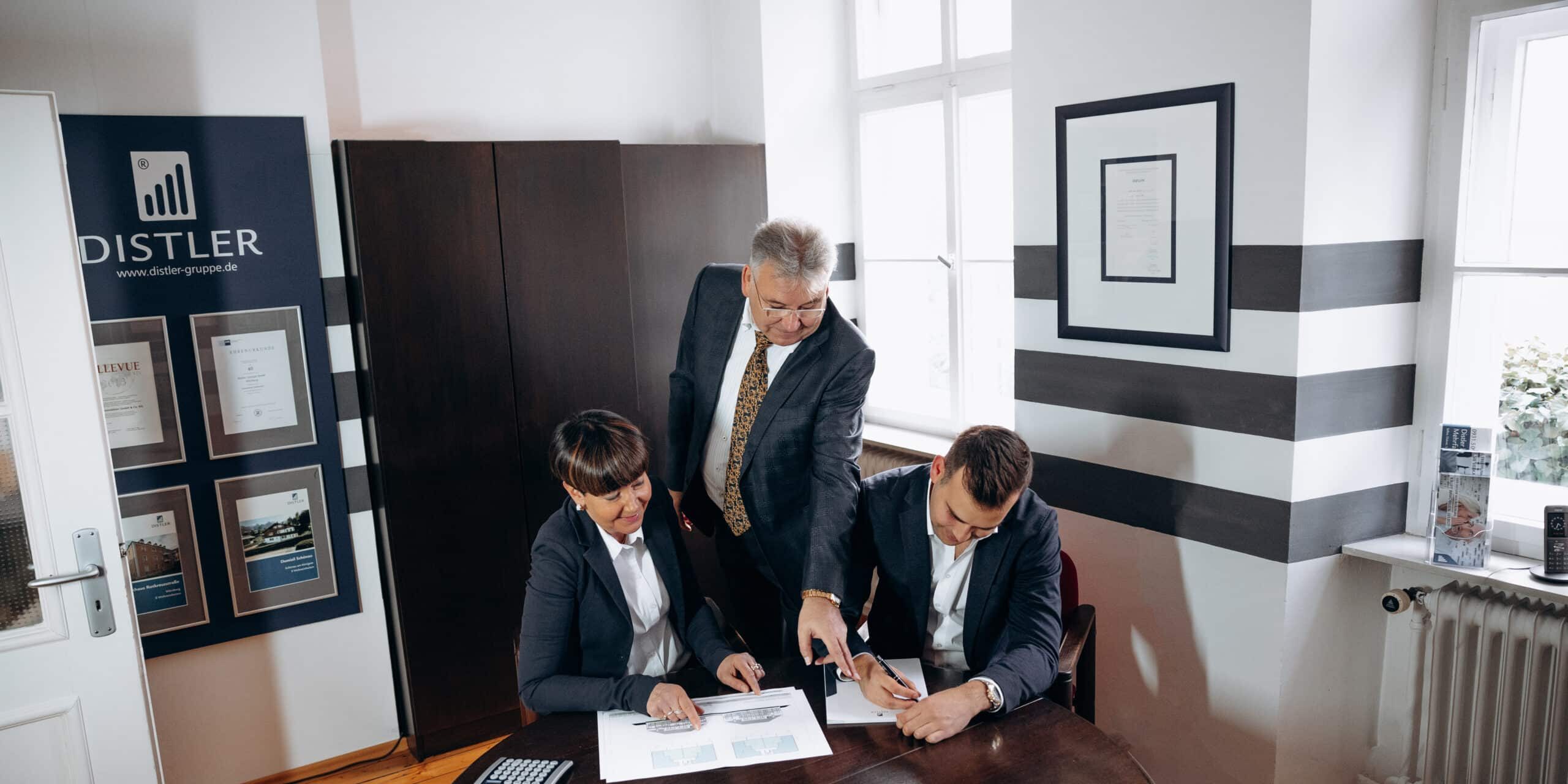 Zwei Männer und eine Frau in Businesskleidung sitzen und stehen an einem dunklen Holztisch, auf dem mehrere Architekturpläne liegen; der stehende Mann zeigt auf einen Punkt auf dem Plan, während die beiden sitzenden Personen Notizen machen und zuhören; im Hintergrund ein Fenster und ein dunkler Schrank.