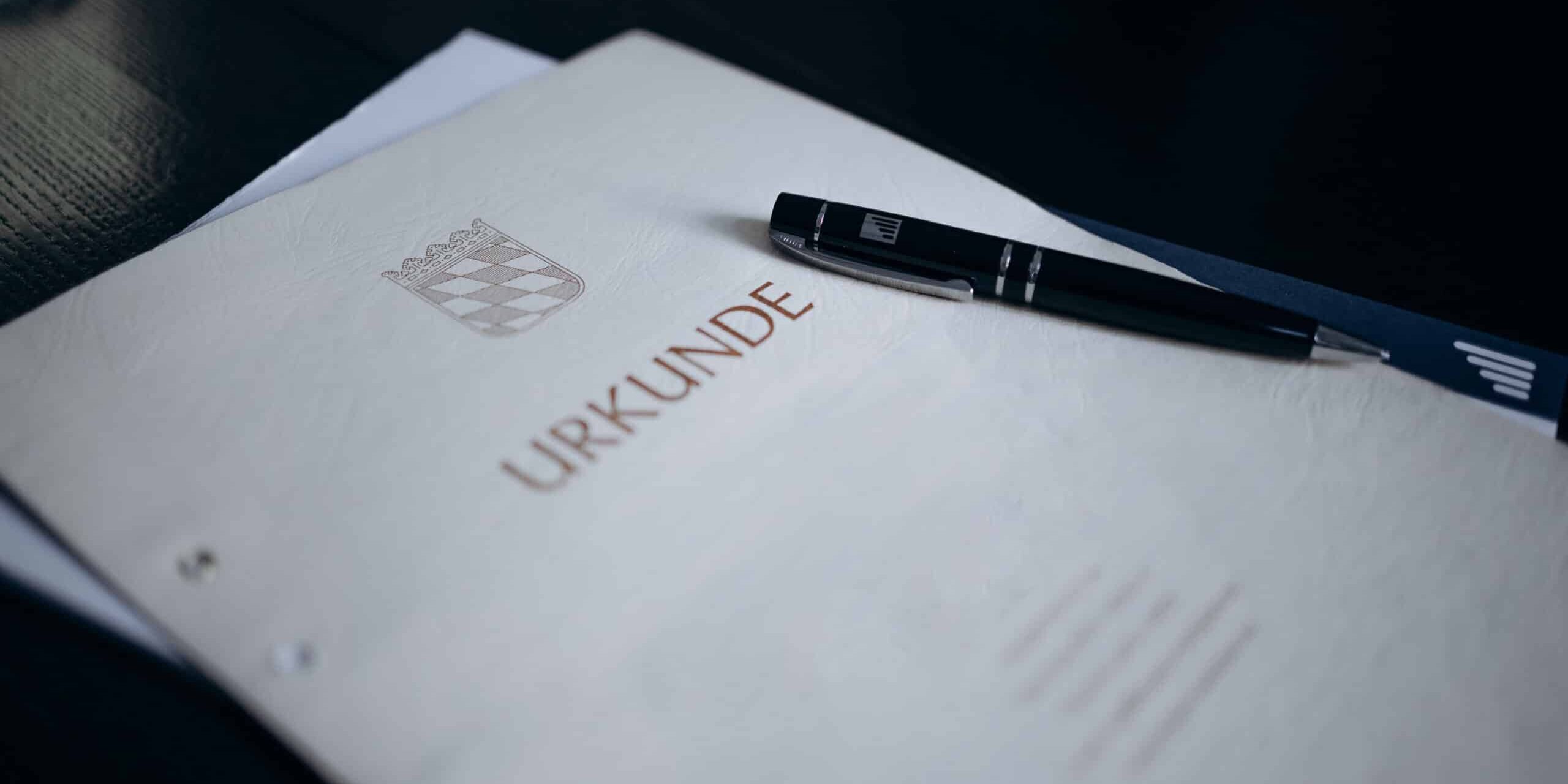 Beige Urkunde mit bayerischem Rautenmuster und der Aufschrift „URKUNDE“ liegt auf einem Stapel Papier; darauf ein schwarzer Kugelschreiber mit silbernem Clip und DISTLER-Aufdruck, Hintergrund ist eine dunkle Tischoberfläche.