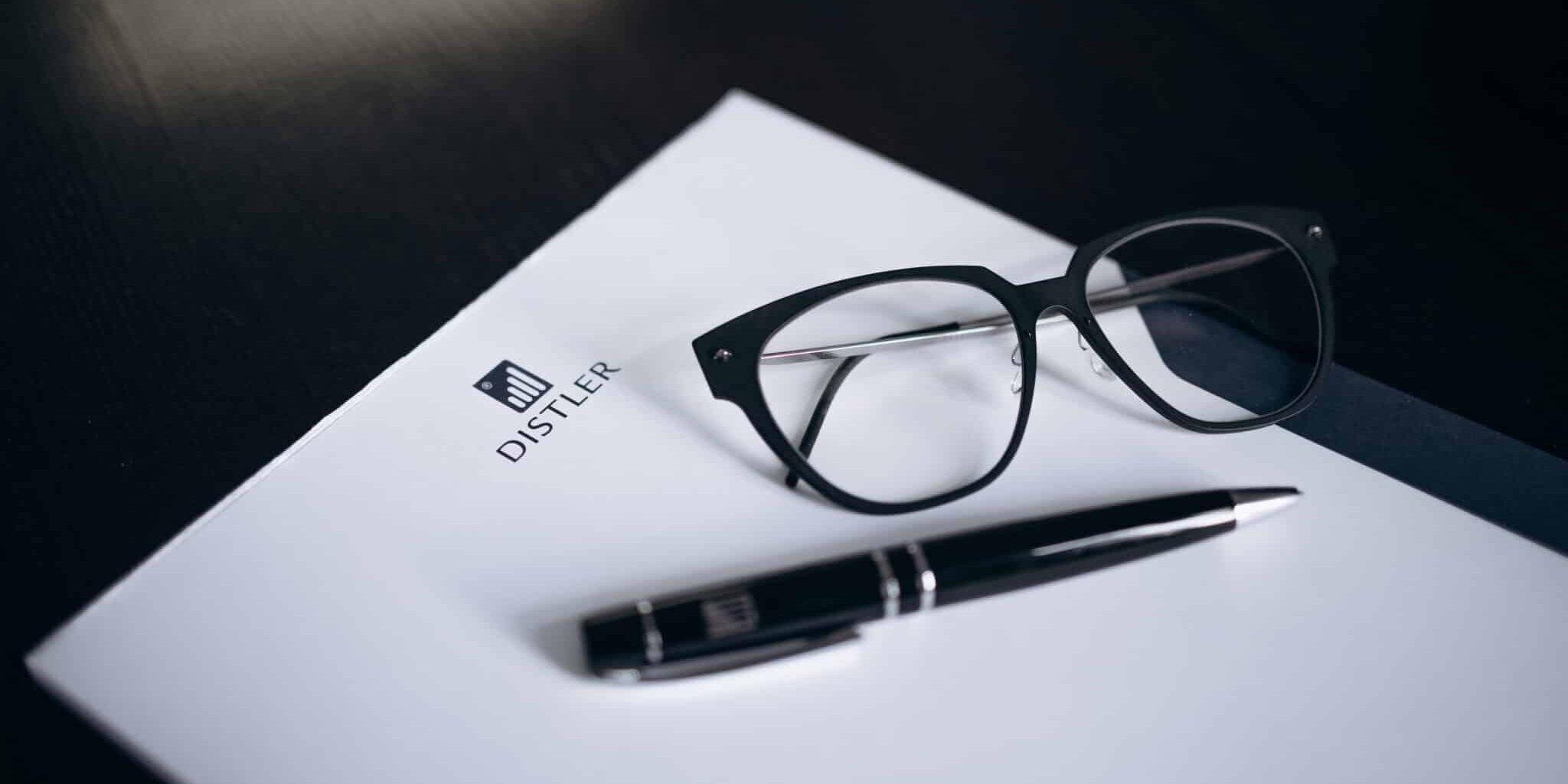 Schwarze Brille und schwarzer Kugelschreiber mit silbernen Details liegen auf einem weißen Schreibblock mit DISTLER-Logo, Hintergrund ist eine dunkle Tischoberfläche.