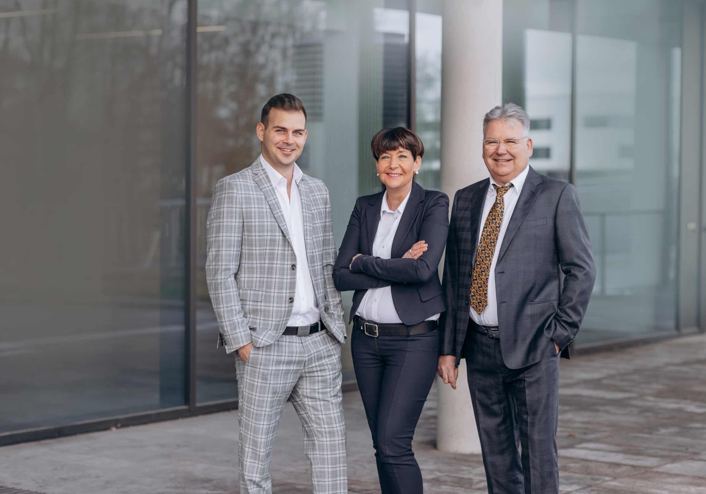 Zwei Männer und eine Frau stehen auf grauem Pflaster vor einem modernen Gebäude mit Fensterfront.