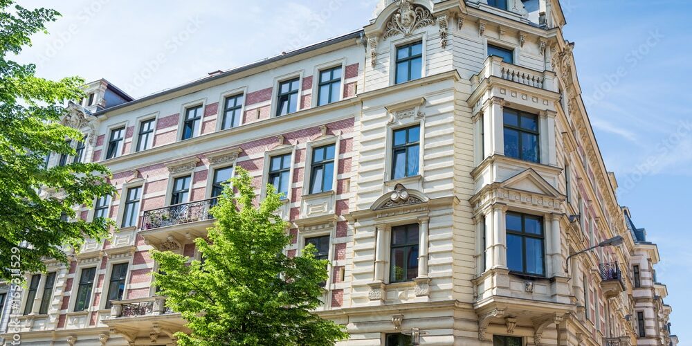 Viergeschossiges Wohn- und Geschäftshaus mit aufwendig verzierter Stuckfassade, Erkern, Balkonen und dekorativen Fensterelementen, im Vordergrund ein grüner Baum.