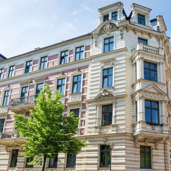 Viergeschossiges Wohn- und Geschäftshaus mit aufwendig verzierter Stuckfassade, Erkern, Balkonen und dekorativen Fensterelementen, im Vordergrund ein grüner Baum.