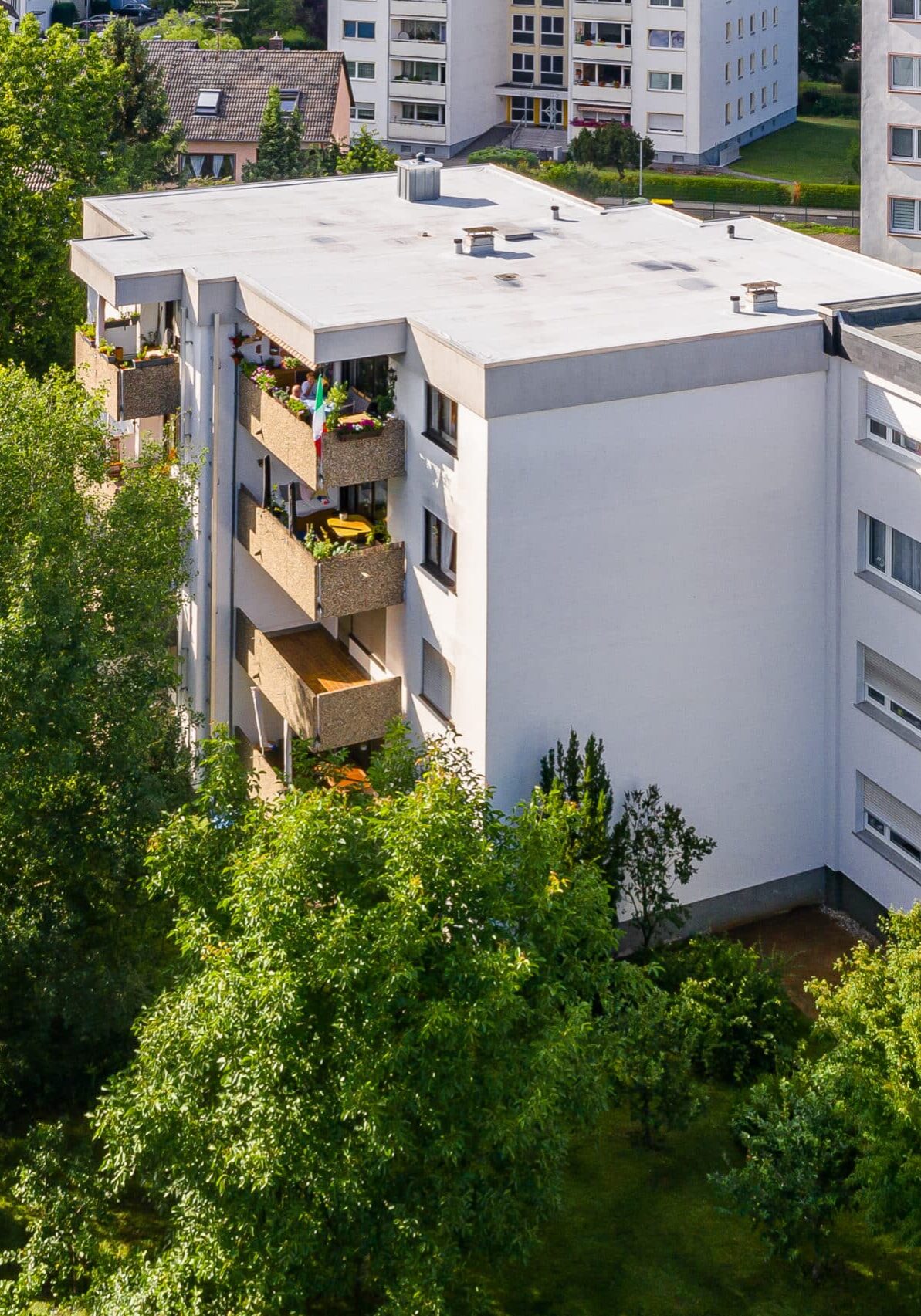 Fünfgeschossiges Wohnhaus mit Flachdach, weißer Putzfassade und mehreren bepflanzten Balkonen, umgeben von Bäumen; im Hintergrund weitere Wohngebäude sichtbar.