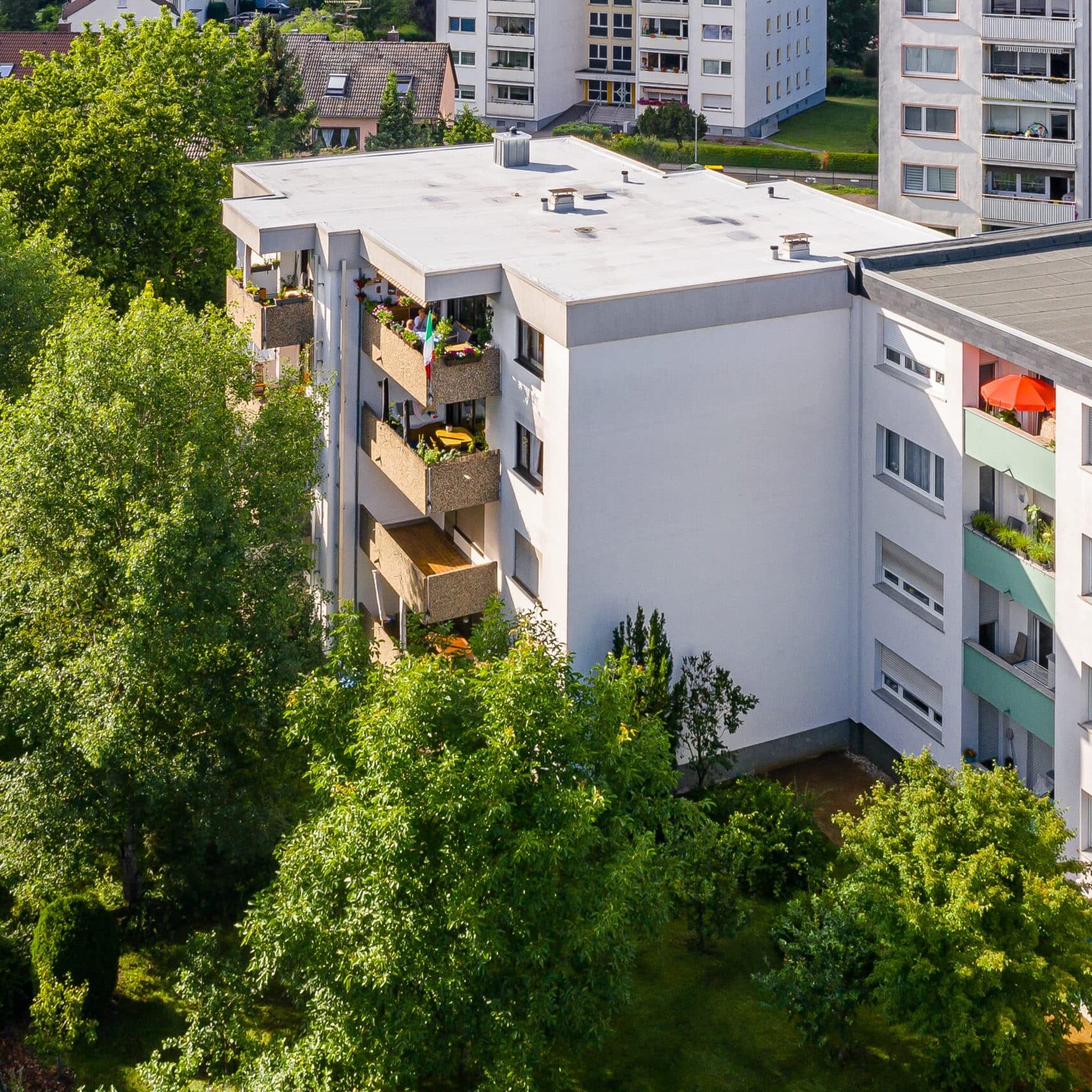Fünfgeschossiges Wohnhaus mit Flachdach, weißer Putzfassade und mehreren bepflanzten Balkonen, umgeben von Bäumen; im Hintergrund weitere Wohngebäude sichtbar.