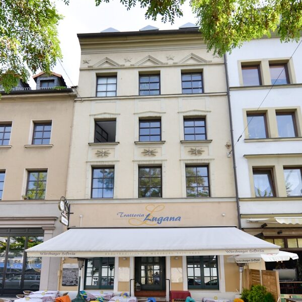 Viergeschossiges Gebäude mit heller Fassade, symmetrischer Fensteranordnung und Markise im Erdgeschoss, darunter ein Restaurant mit Außengastronomie und weißen Tischen.