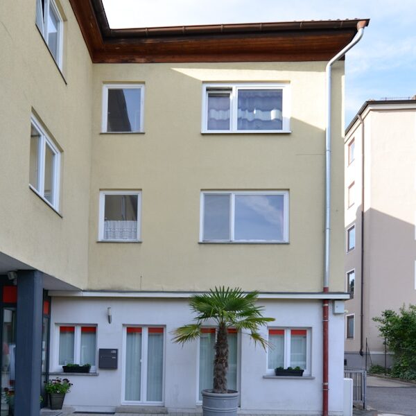 Dreigeschossiges Wohnhaus mit gelber Fassade, weißen Fenstern, kleinem Innenhofbereich und zwei Kübelpalmen vor dem Eingang im Erdgeschoss.