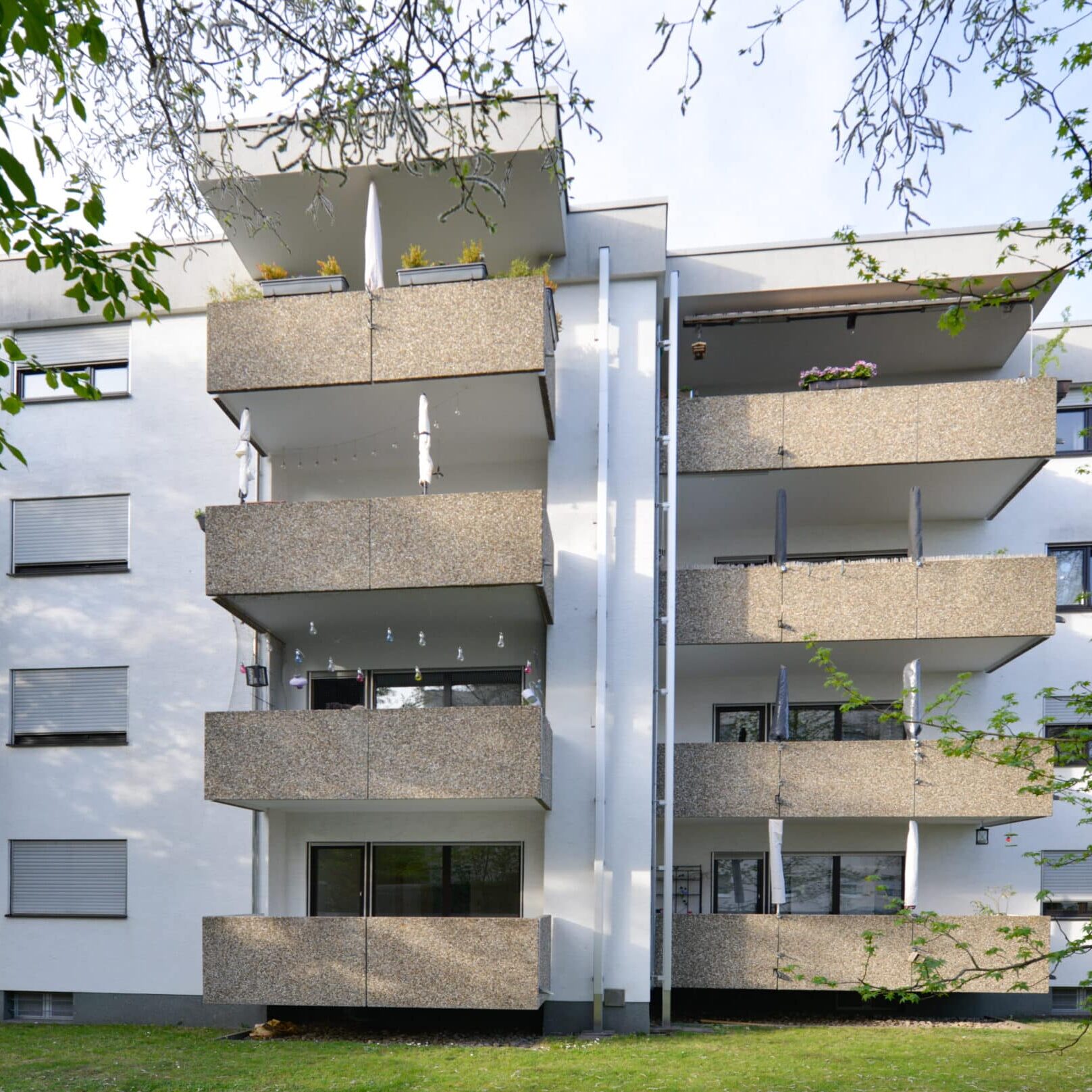Mehrgeschossiges Wohngebäude mit außenliegenden Betonbalkonen, weißer Fassade, teils verdeckt durch Bäume im Vordergrund.