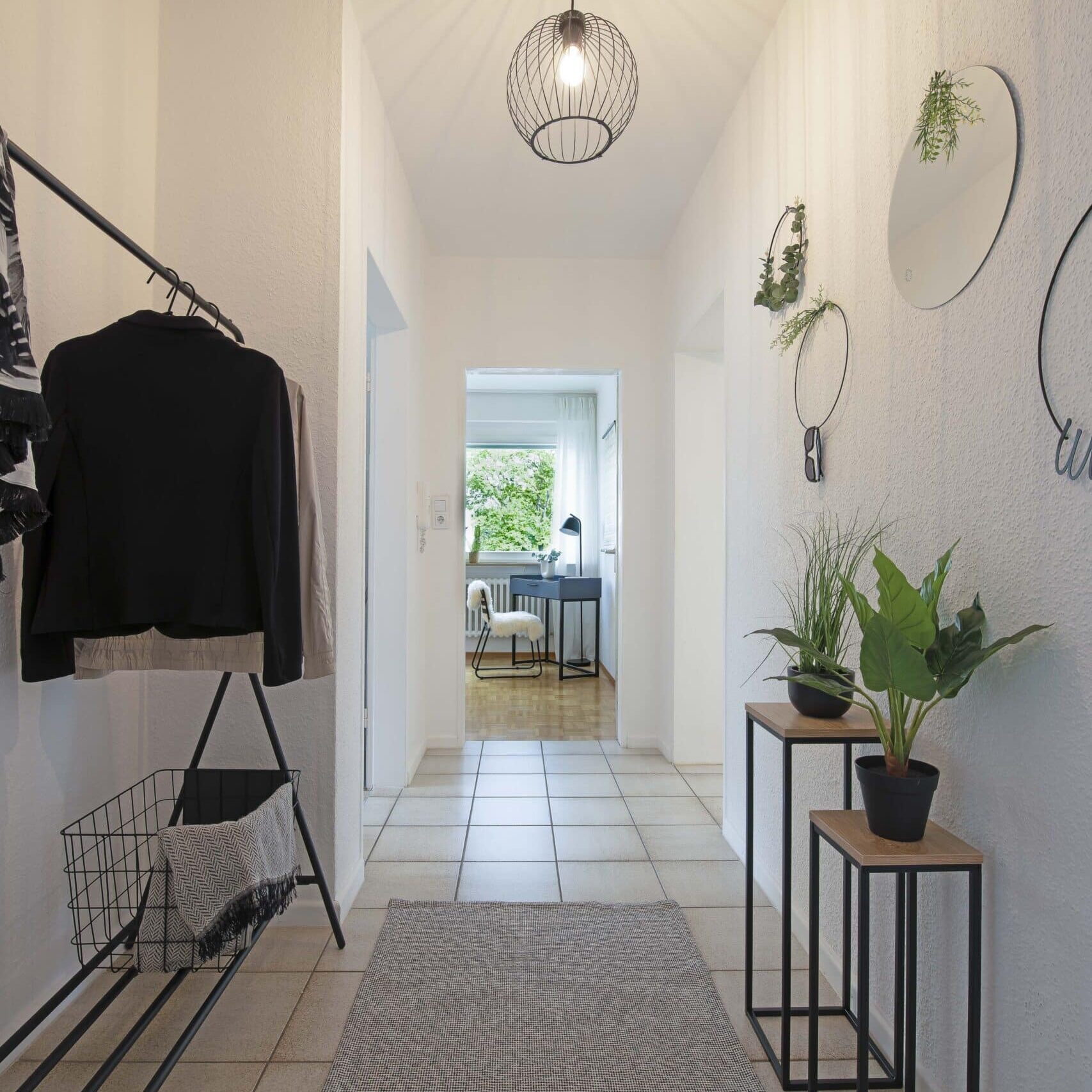 Langer Flur mit Fliesenboden, hellen Wänden, Pflanzen und runden Spiegeln an der rechten Wand, im Hintergrund ein Wohnraum mit Tageslicht.