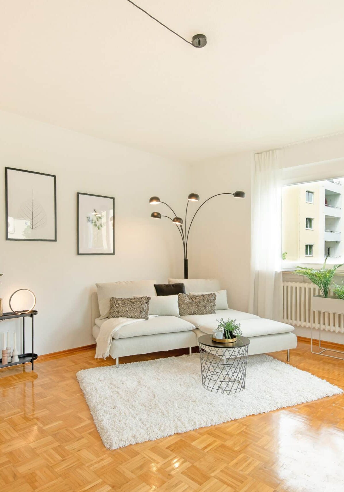 Modern eingerichtetes Wohnzimmer mit glänzendem Parkettboden, weißem Sofa, Teppich, runder Couchtisch, Bogenlampe, Pflanzen am Fensterbrett und Blick auf Nachbargebäude durch das Fenster.