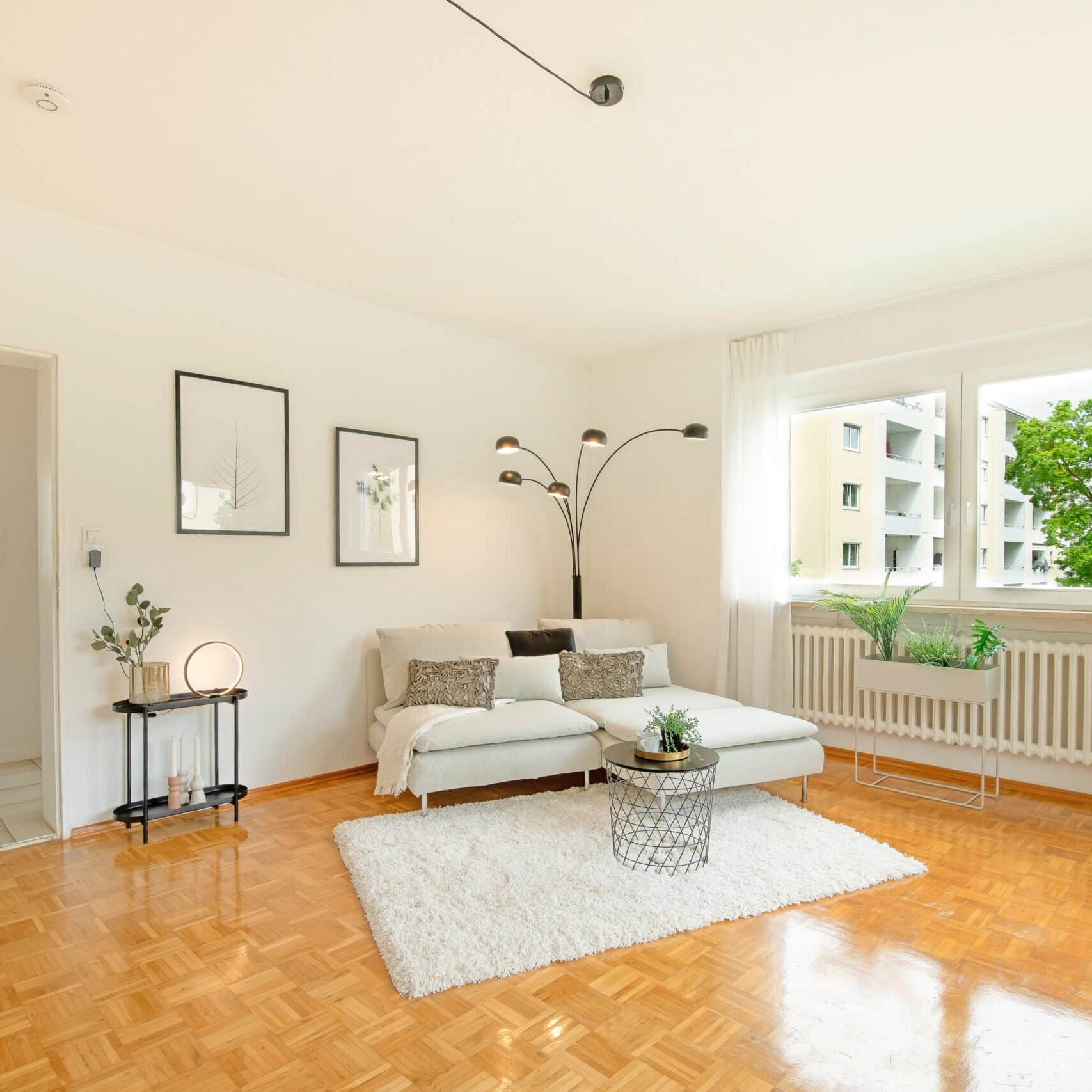Modern eingerichtetes Wohnzimmer mit glänzendem Parkettboden, weißem Sofa, Teppich, runder Couchtisch, Bogenlampe, Pflanzen am Fensterbrett und Blick auf Nachbargebäude durch das Fenster.