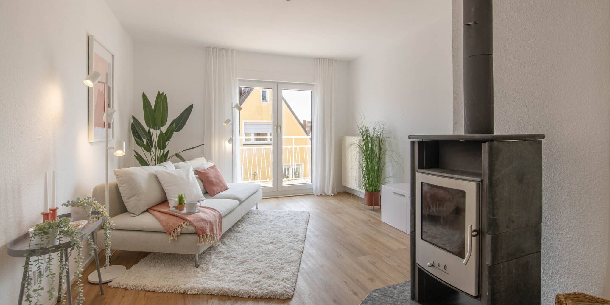 Modern eingerichtetes Wohnzimmer mit hellem Laminatboden, weißem Sofa mit Kissen und Decke, flauschigem Teppich, bodentiefen Fenstern mit Balkonzugang, Vorhängen, mehreren Zimmerpflanzen, einem Kaminofen im Vordergrund rechts und Tageslicht aus südlicher Richtung.
