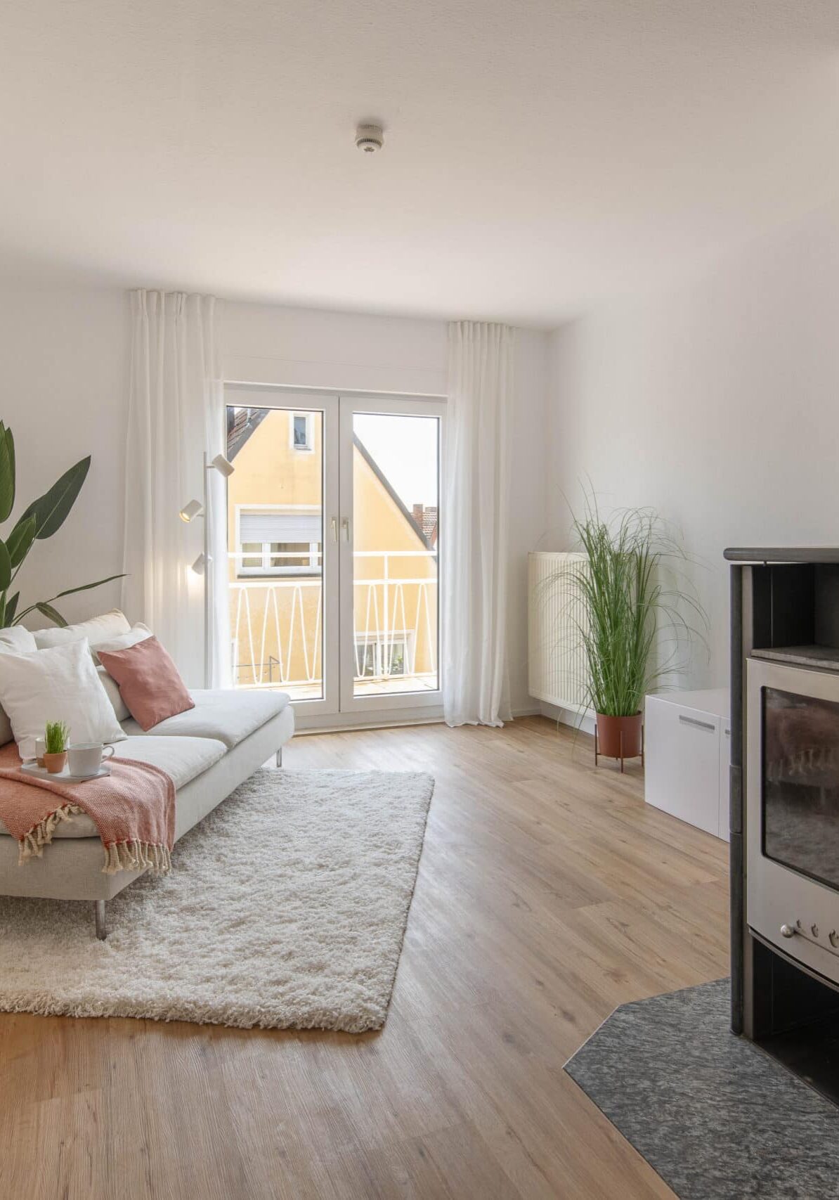 Modern eingerichtetes Wohnzimmer mit hellem Laminatboden, weißem Sofa mit Kissen und Decke, flauschigem Teppich, bodentiefen Fenstern mit Balkonzugang, Vorhängen, mehreren Zimmerpflanzen, einem Kaminofen im Vordergrund rechts und Tageslicht aus südlicher Richtung.