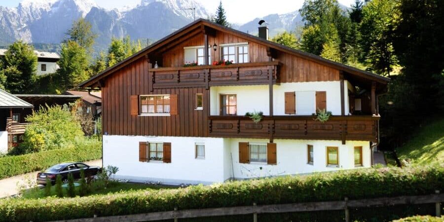 Zweigeschossiges Mehrfamilienhaus mit Holzfassade und Satteldach, Balkon und Fensterläden, im Vordergrund ein Holzzaun; Hintergrund zeigt bewaldete Berge mit Schnee.