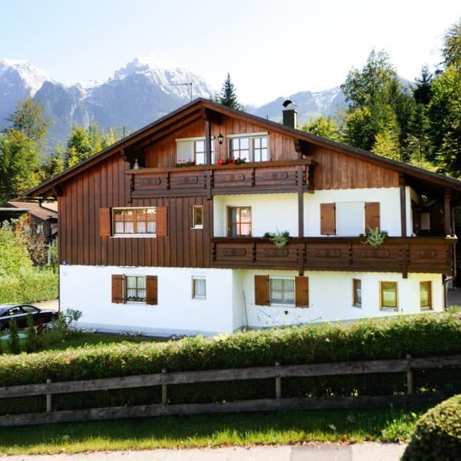 Zweigeschossiges Mehrfamilienhaus mit Holzfassade und Satteldach, Balkon und Fensterläden, im Vordergrund ein Holzzaun; Hintergrund zeigt bewaldete Berge mit Schnee.