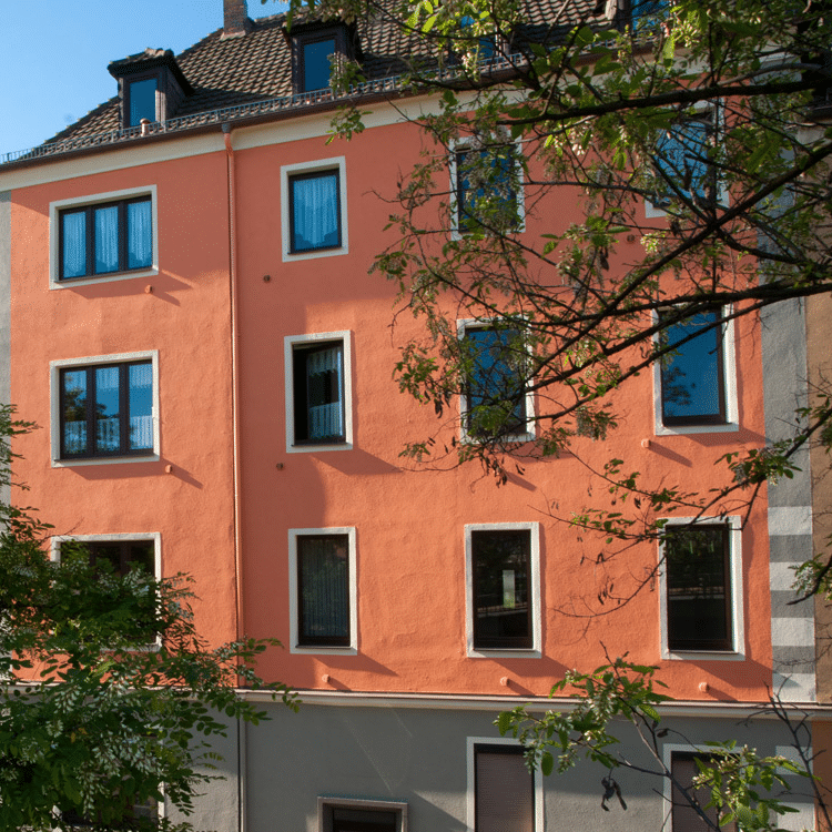 Viergeschossiges Wohnhaus mit orangefarbener Putzfassade, weißen Fensterrahmen und dunklem Dach, teils verdeckt durch Laubbäume im Vordergrund.