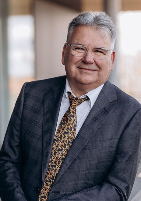 Ein Mann mit grauen Haaren, Brille, dunklem karierten Anzug, weißem Hemd und gold-braun gemusterter Krawatte steht leicht schräg und lächelt; im Hintergrund unscharfe Gebäudeelemente mit Licht.