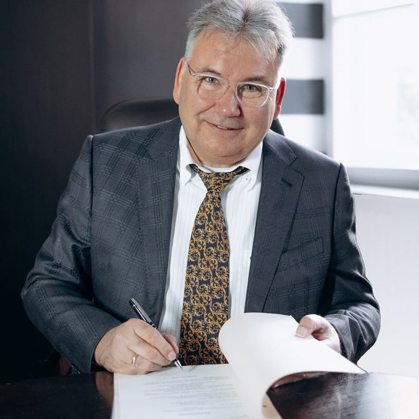 Mann mit grauen Haaren, Brille, gestreiftem Hemd, kariertem Sakko und gemusterter Krawatte sitzt an einem dunklen Tisch, hält einen Stift und ein geöffnetes Dokument.