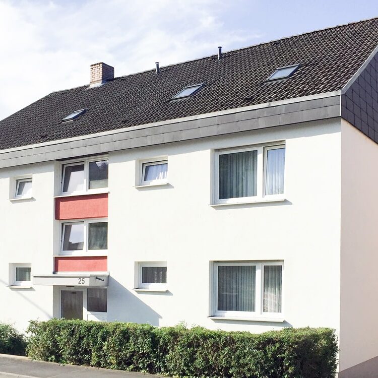 Dreigeschossiges Mehrfamilienhaus mit weißer Fassade, roten Akzenten, Satteldach und kleinen Fenstern, davor ein schmaler Vorgarten mit Hecke.