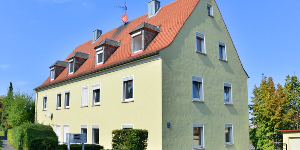 Dreigeschossiges Wohngebäude mit hellgelber Putzfassade, weißen Fensterrahmen und rotem Satteldach, davor ein niedriger grüner Strauch.