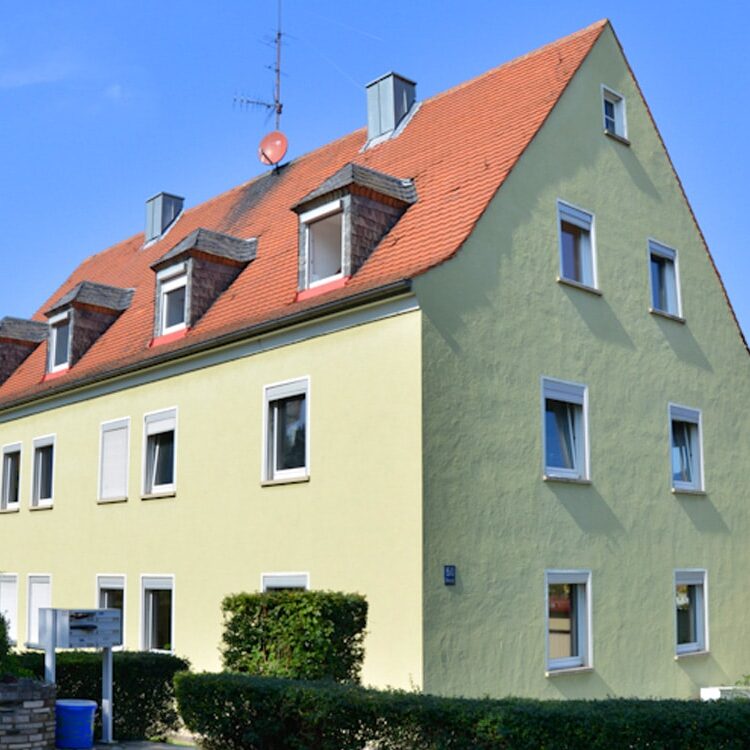 Dreigeschossiges Wohngebäude mit hellgelber Putzfassade, weißen Fensterrahmen und rotem Satteldach, davor ein niedriger grüner Strauch.