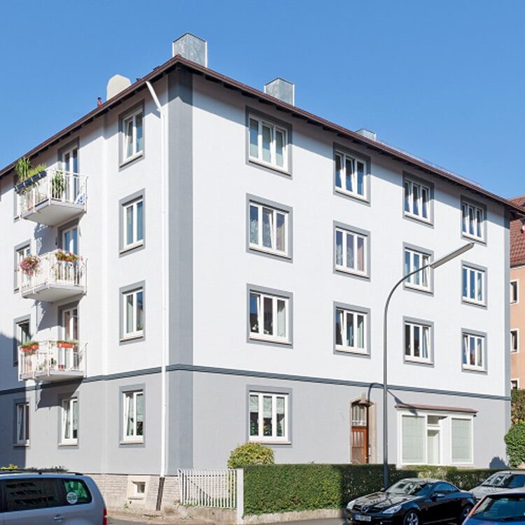 Helles, viergeschossiges Wohnhaus mit mehreren Balkonen, weißen Fenstern und hellgrauer Sockelzone, davor Gehweg und geparkte Fahrzeuge.
