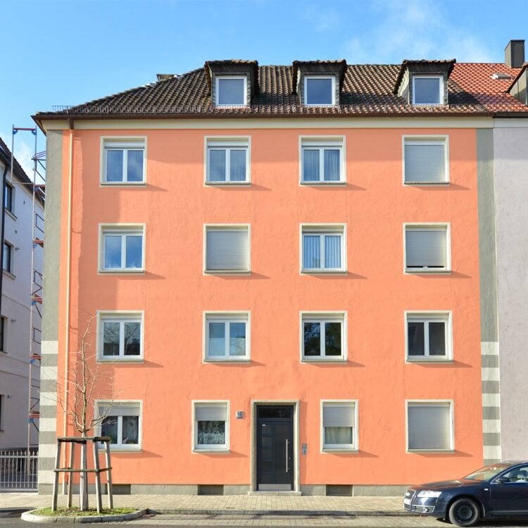 Fünfgeschossiges Wohnhaus mit orangefarbener Putzfassade, weißen Fensterrahmen, dunklem Satteldach und geparktem Auto im Vordergrund.