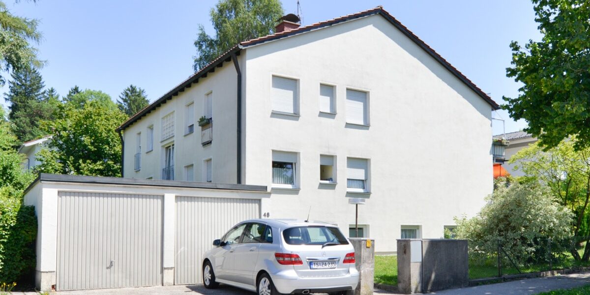 Rückansicht eines gepflegten Mehrfamilienhauses mit weißer Fassade, Garage, Parkplatz und Grünanlage.