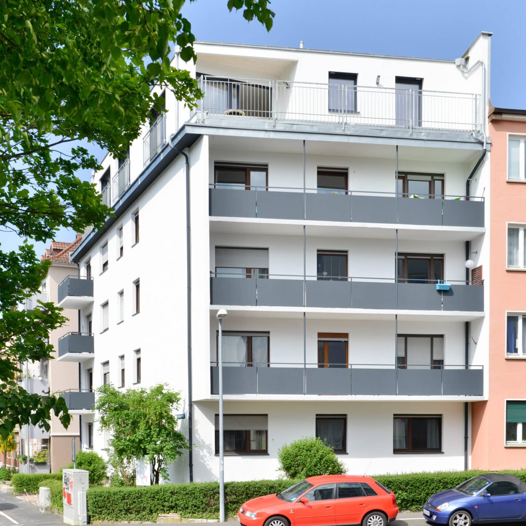 Fünfgeschossiges weißes Wohngebäude mit durchgehenden Balkonen, Flachdach, grauen Brüstungen und parkenden Autos im Vordergrund.