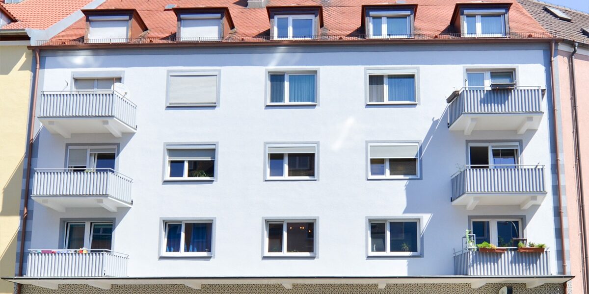 Viergeschossiges Gebäude mit weißer Fassade, mehreren Fenstern, einem Ladengeschäft im Erdgeschoss und Balkonen zur Straße.